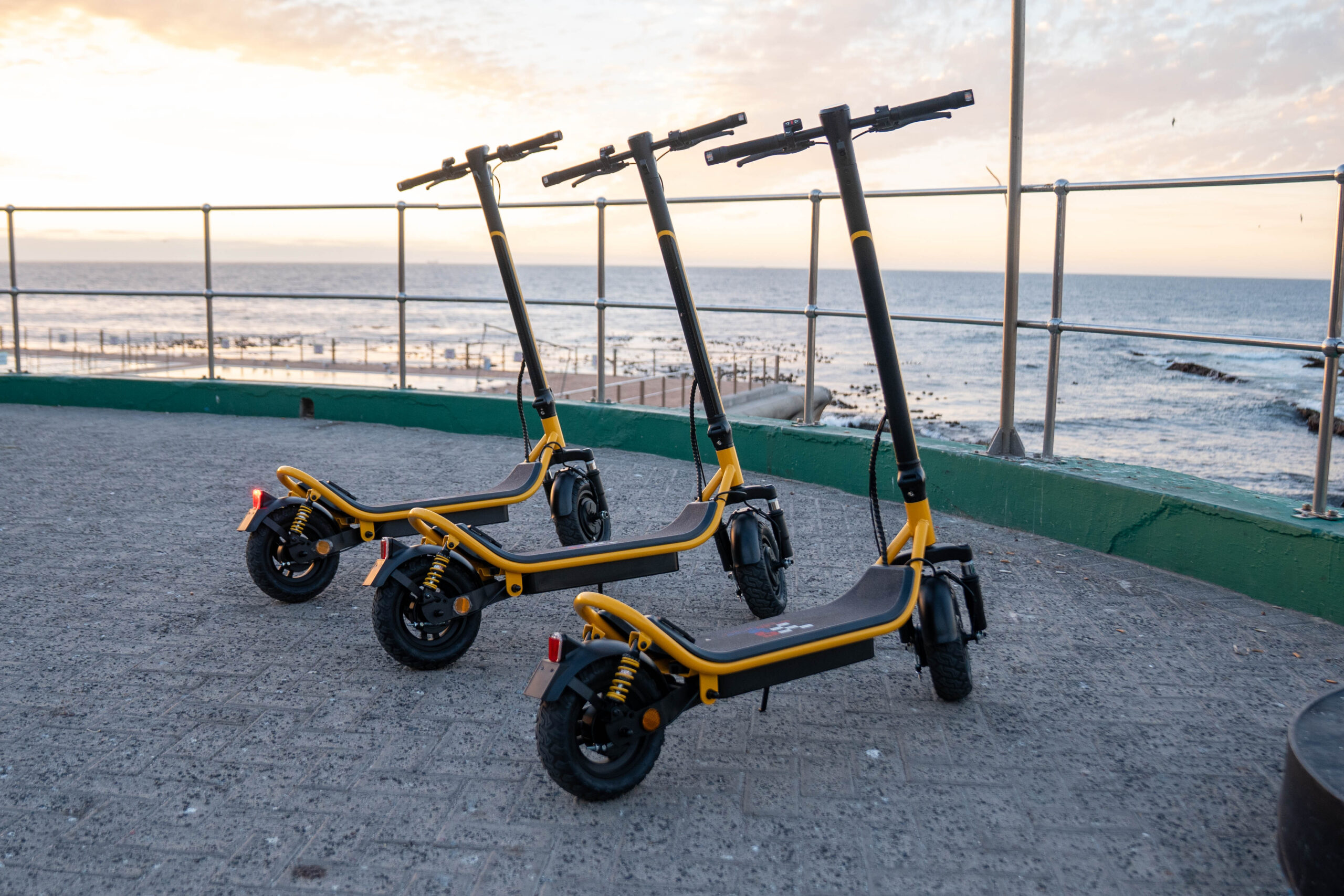 Three yellow and black Speedbikes electric scooters lined up on a seafront promenade at sunset